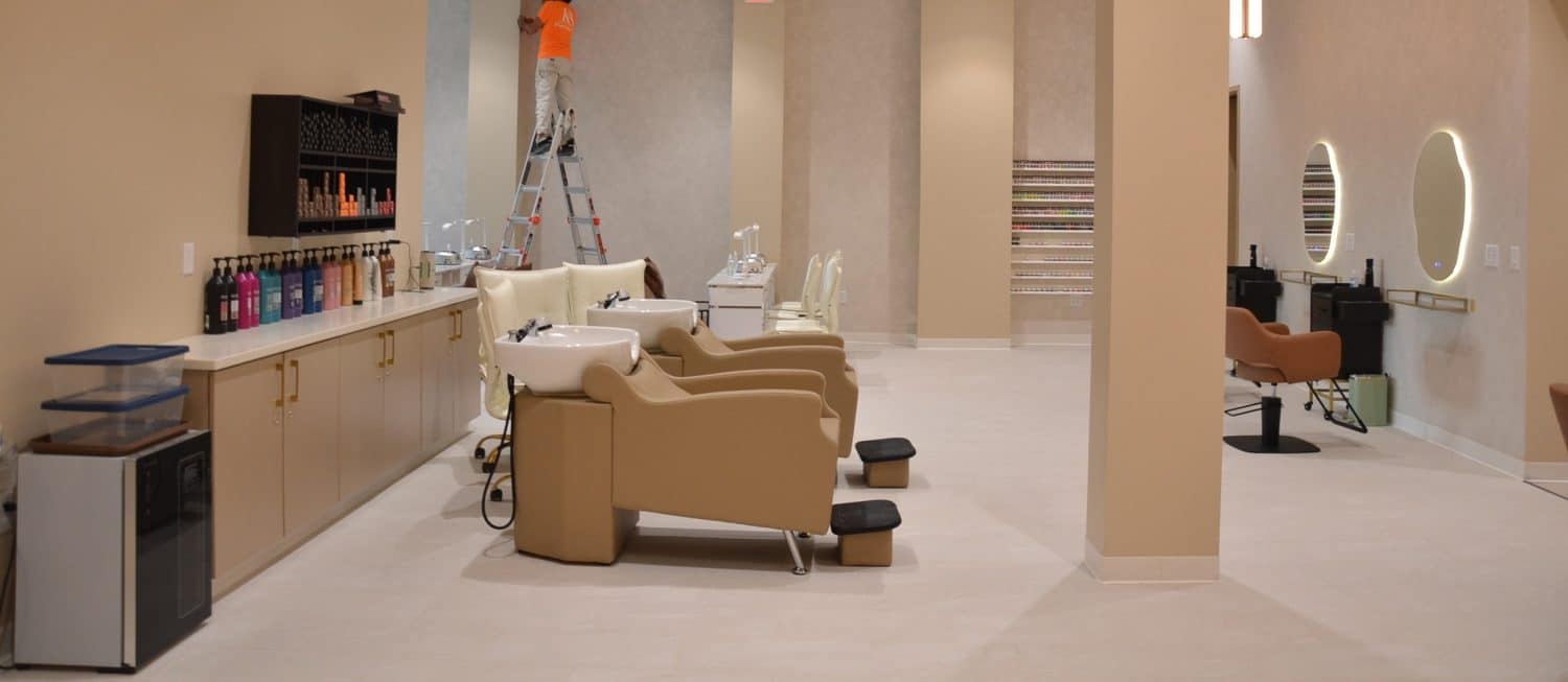 A modern salon interior with beige chairs, washing stations, product shelves, and a worker on a ladder performing maintenance on the back wall.