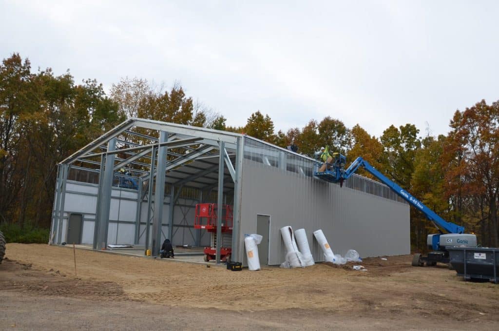 Workers use a lift to install panels on a steel-framed building under construction, with construction materials on the ground and trees in the background.