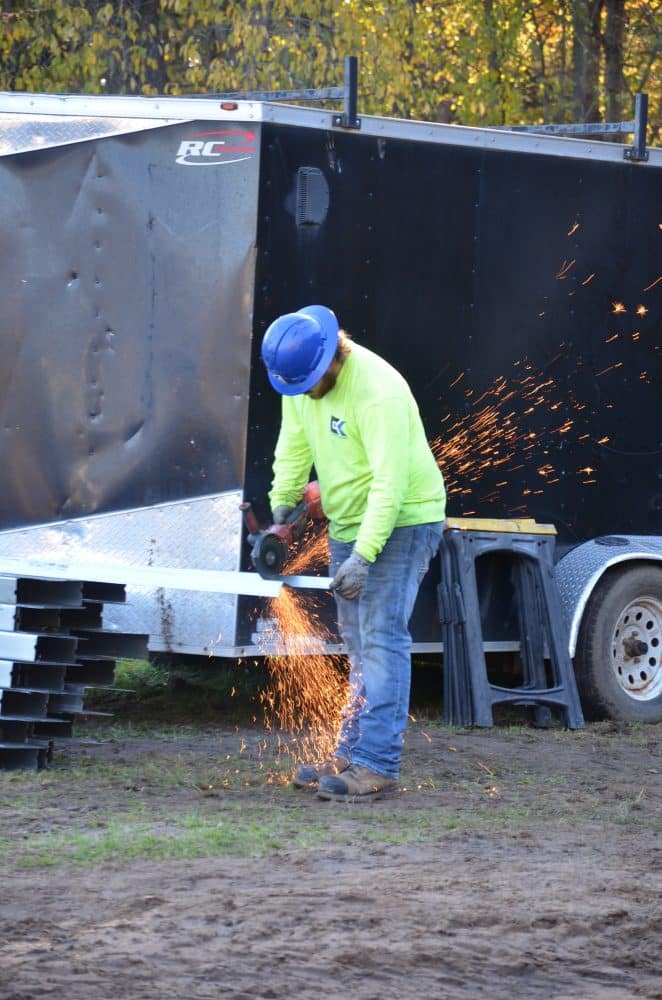 A construction worker in a yellow shirt and blue helmet uses a power tool to cut metal, producing sparks, near a trailer and stacks of materials outdoors.