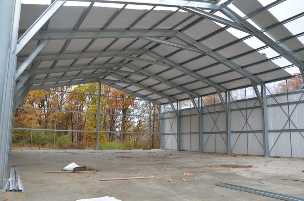 Interior view of a partially constructed metal building with an open end, exposed steel framework, and trees visible outside.