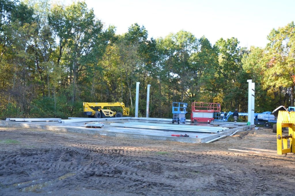 Construction site with steel beams and columns being erected, several vehicles and equipment present, surrounded by trees and dirt ground.