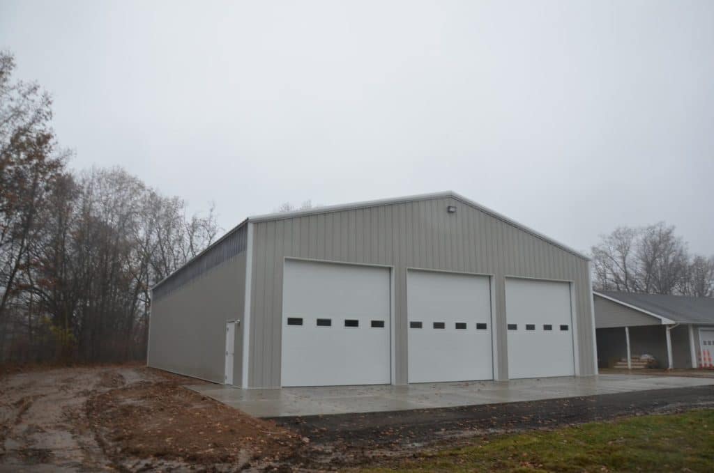 A large, light gray metal building with three white garage doors sits on a concrete pad, surrounded by trees and overcast sky.