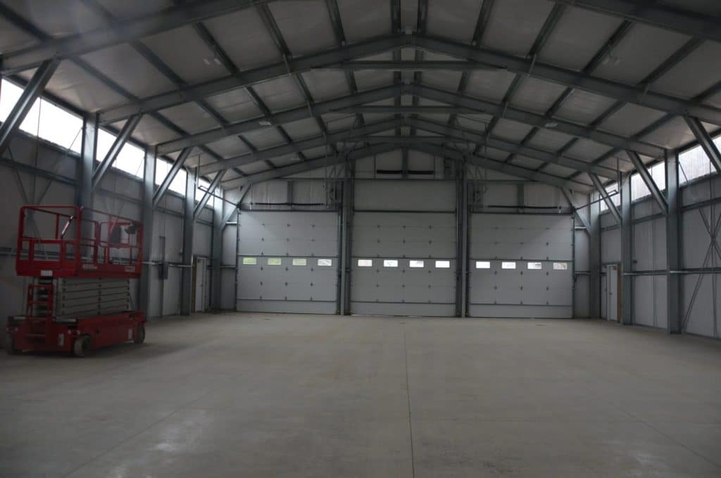 Interior of a large empty warehouse with concrete floors, steel beams, multiple garage doors, and a red scissor lift parked on the left.