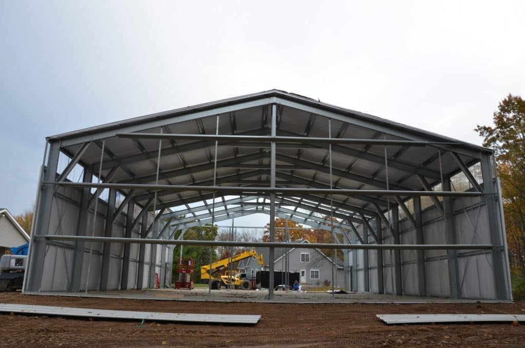 A large steel-framed building under construction, with a crane and construction equipment inside, and trees and a house in the background.