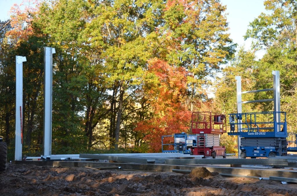 Steel beams and columns being erected at a construction site, with scissor lifts nearby and trees with autumn foliage in the background.