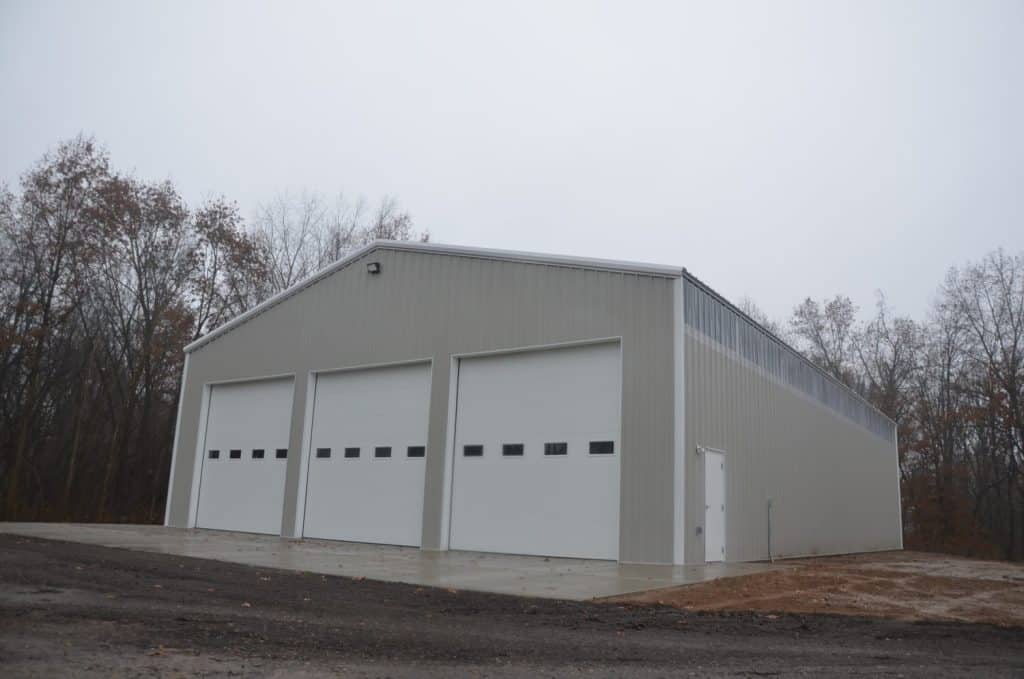 A large, beige metal building with three garage doors and a side entrance door, situated on a concrete pad near a wooded area under an overcast sky.
