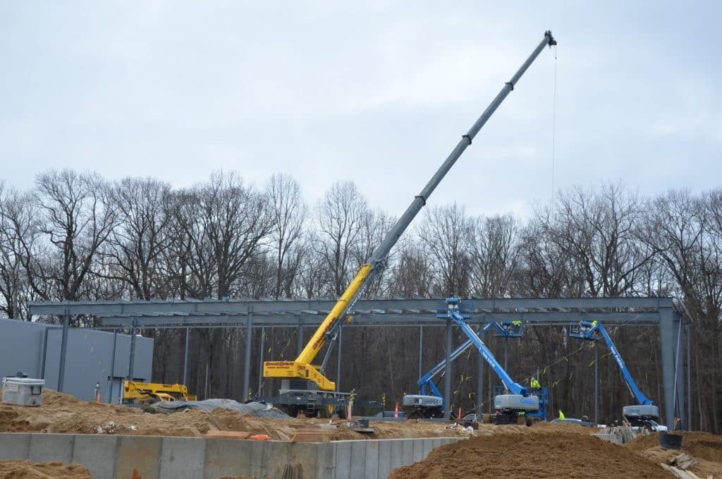 A construction site with cranes and workers assembling a steel framework for a building, surrounded by dirt piles and bare trees in the background.