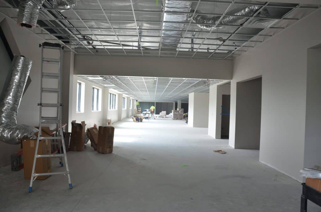 A large indoor commercial space under construction with exposed ceiling ducts, a ladder, boxes, and workers visible in the background.