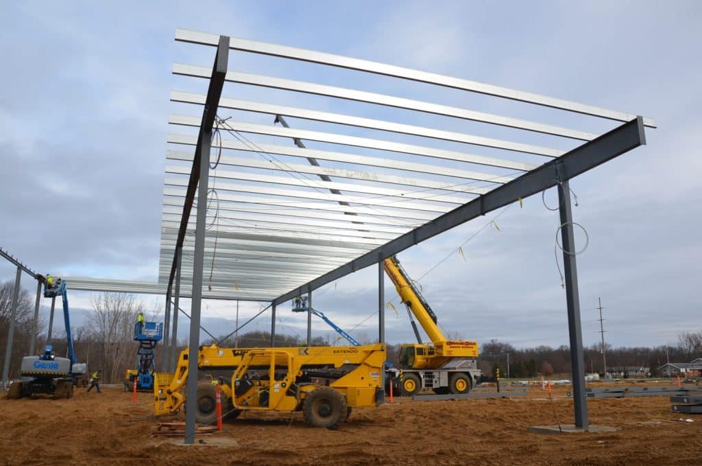 Construction site with steel framework being assembled; workers operate yellow cranes and lifts on bare ground under a cloudy sky.