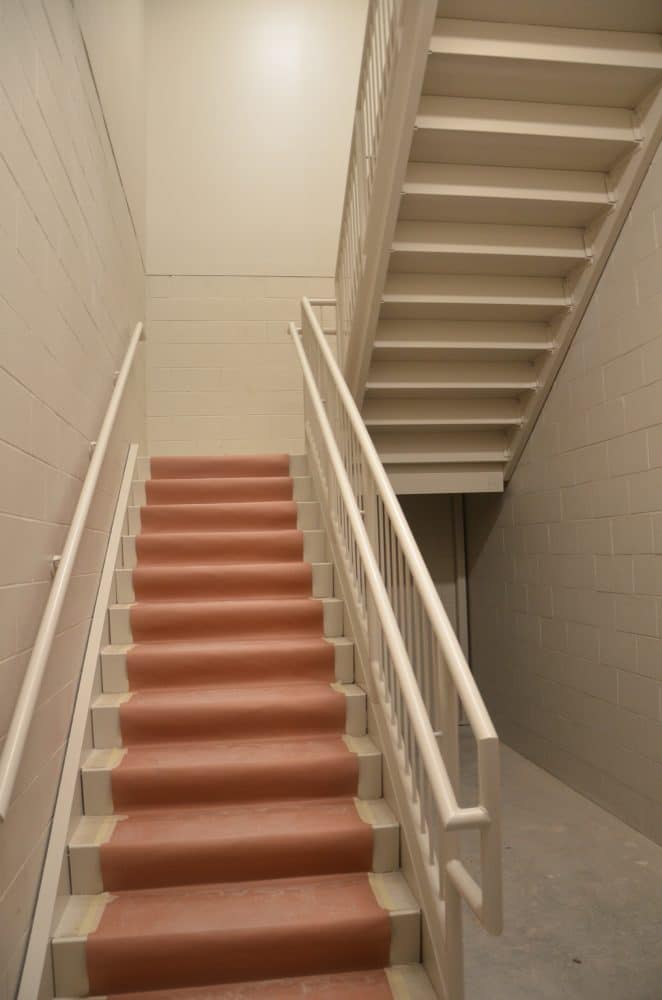 Indoor stairwell with red steps, white railings, and white brick walls, leading to a landing and another flight of stairs above.