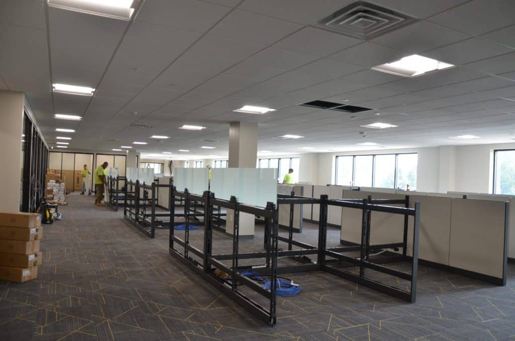 Office space under construction with workers assembling cubicles, boxes stacked on the floor, and ceiling panels partially removed.