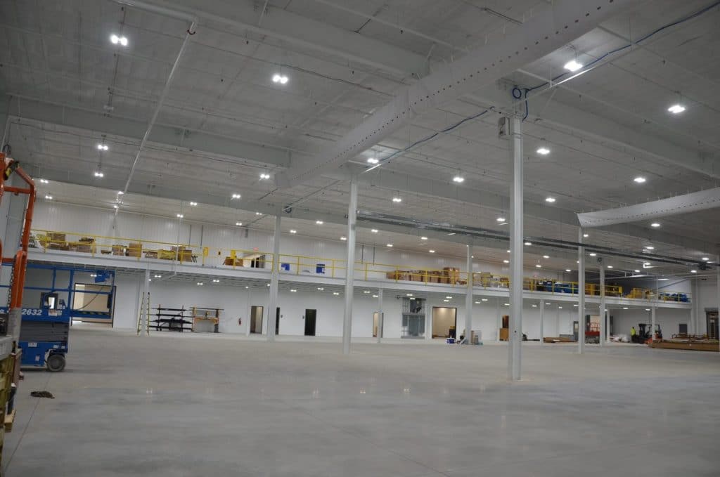 Spacious, well-lit industrial warehouse interior with high ceilings, concrete floor, mezzanine level, and scattered equipment.