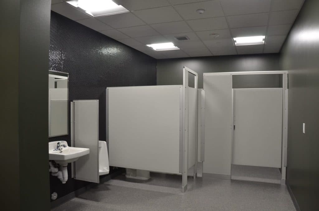 A public restroom with a sink, mirror, urinal, and two toilet stalls with light gray partitions under fluorescent ceiling lights.