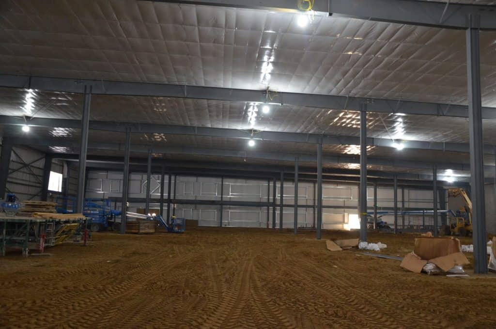 Interior of a large industrial building under construction, with exposed steel beams, equipment, dirt floor, and scattered construction materials.