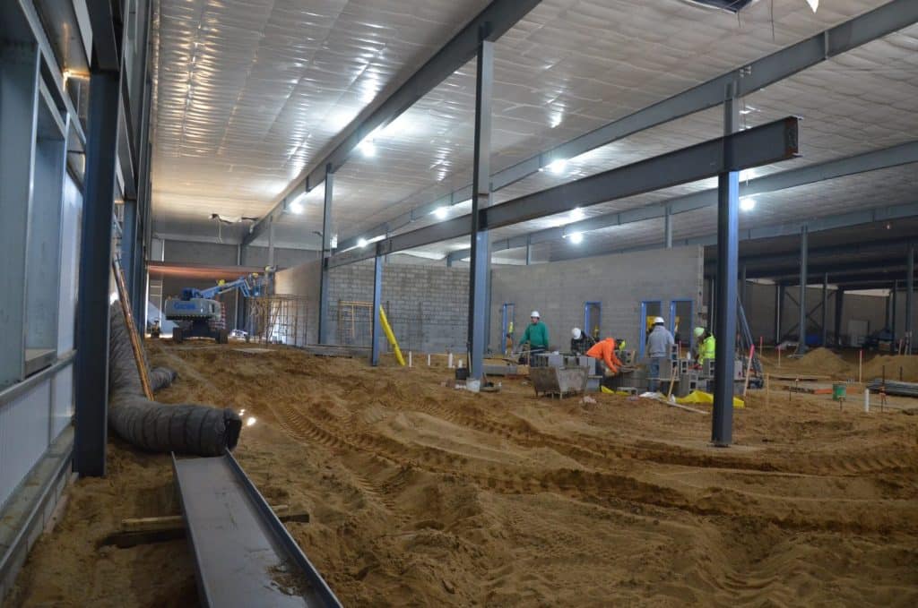 Construction workers in hard hats work inside a large building with exposed beams, unfinished walls, and sandy ground. Equipment and materials are scattered throughout the site.