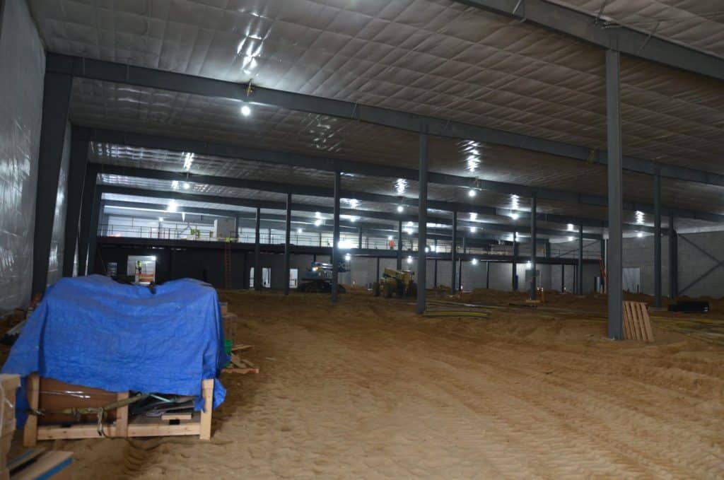 Interior of a large unfinished warehouse with sandy ground, exposed beams, construction materials, and overhead lights.
