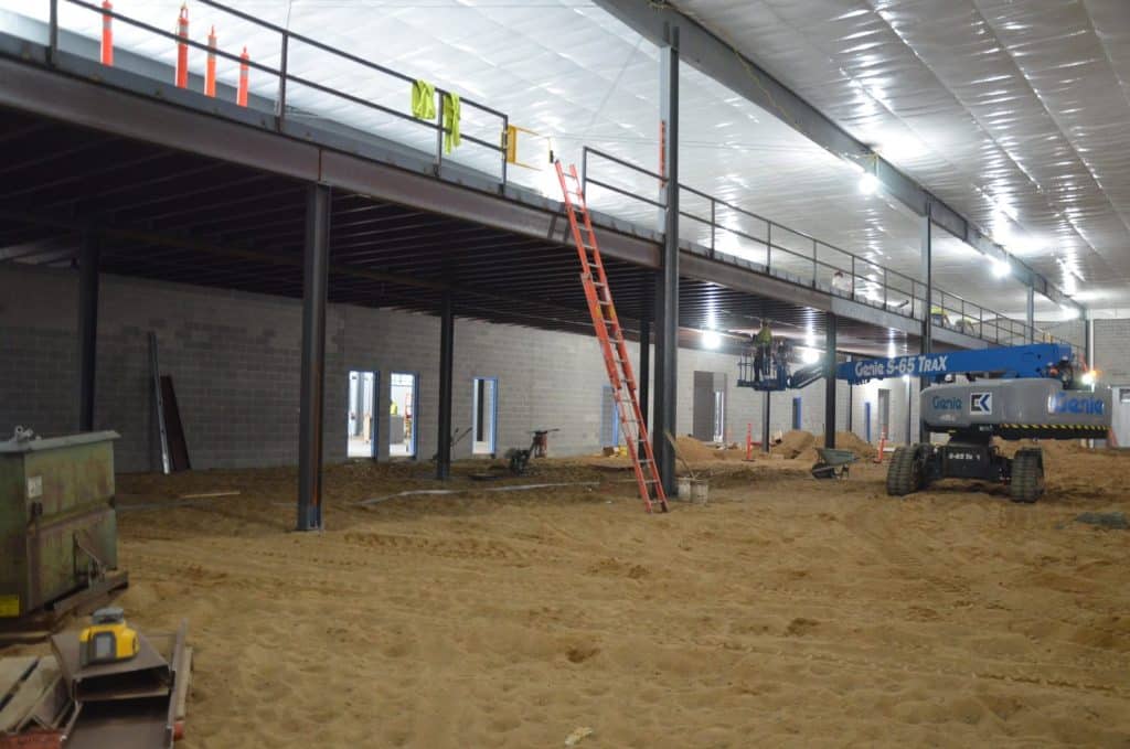 Interior construction site with unfinished walls, exposed beams, a raised platform, a red ladder, and construction equipment on a dirt floor.