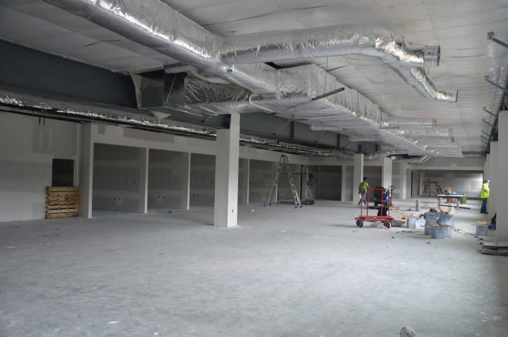 Large indoor space under construction with exposed ductwork, unfinished walls, ladders, construction materials, and several workers in safety vests.