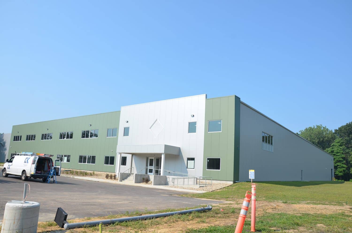 A large, modern industrial building with green and white exterior panels, situated on a grassy lot under a clear blue sky. A white van is parked near the entrance.