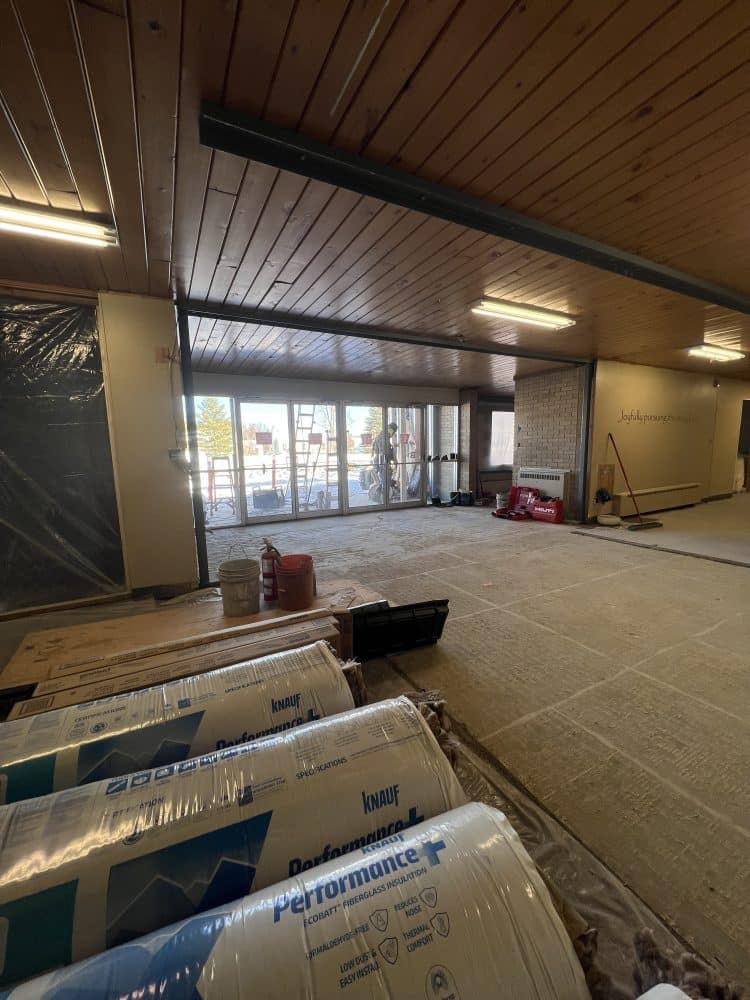 Construction site indoors with insulation rolls, tools, and materials scattered; large windows at the far end show workers and an outdoor area beyond.