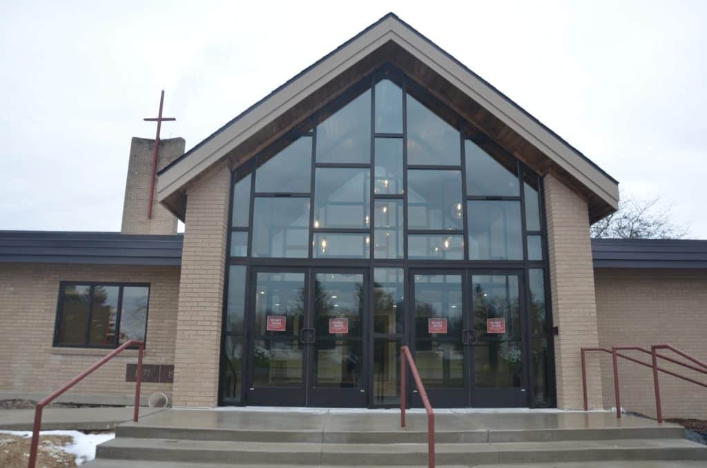 A brick church building with large glass entry doors and windows, a red cross on the chimney, and red railings along the front steps.