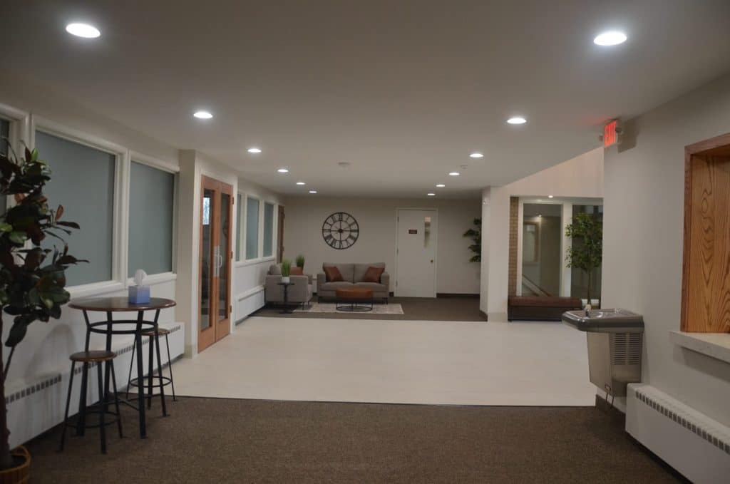Modern hallway with seating area, round wall clock, potted plants, water fountain, and bar-style table with two stools under ceiling lights.