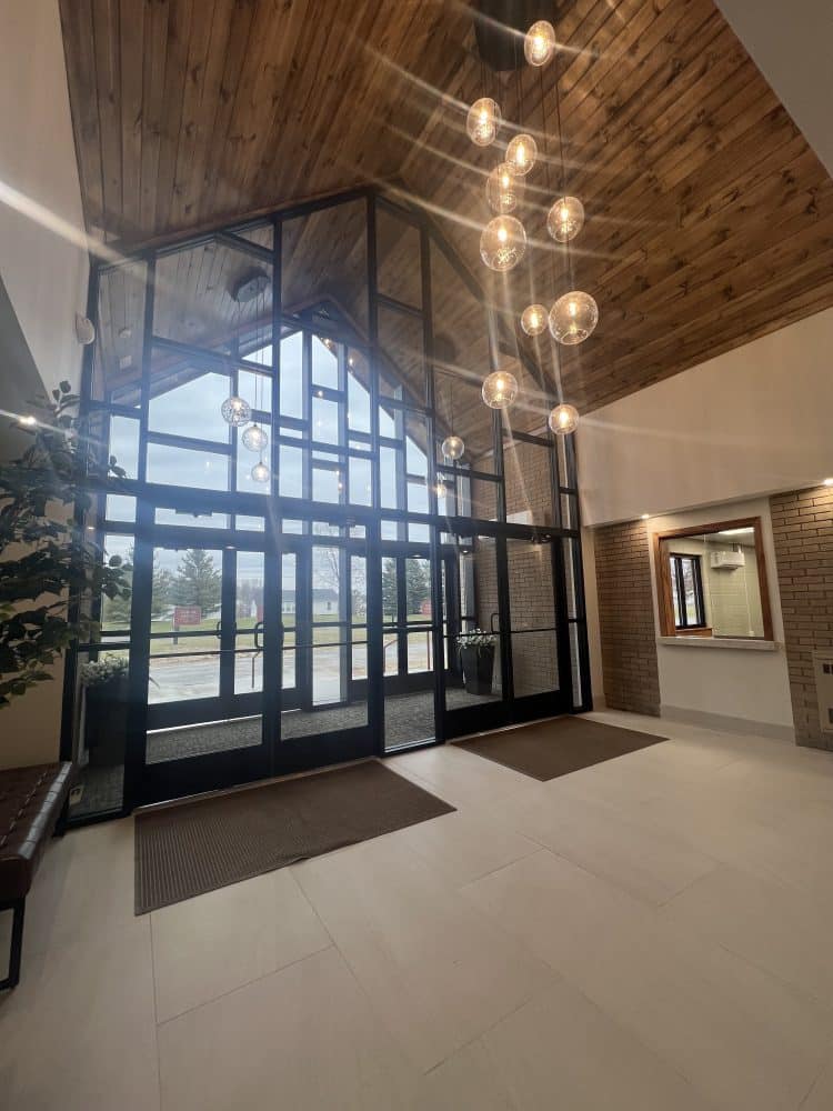 A modern building lobby with large glass windows, a wooden vaulted ceiling, hanging globe lights, and light tile flooring.