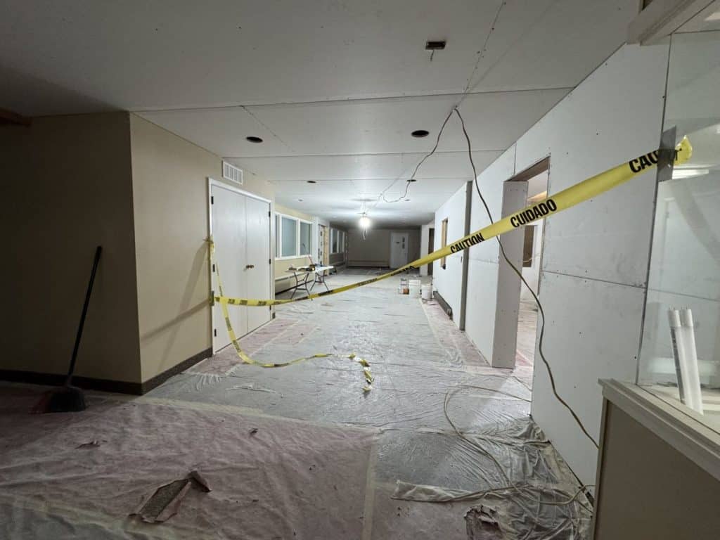 A hallway under construction with exposed wires, unfinished walls, construction materials, and caution tape blocking access.