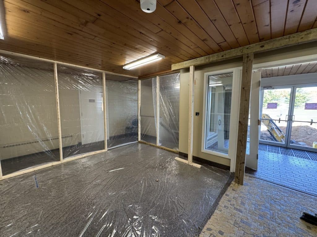 A room under renovation with plastic sheeting covering the floor and an unfinished wall, next to glass doors leading outside.