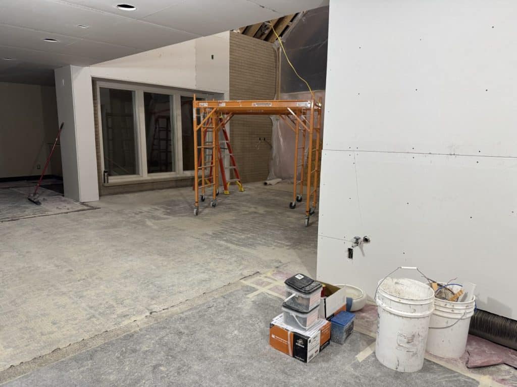 Interior construction site with scaffolding, ladders, exposed drywall, construction materials, and tools scattered on the unfinished floor.