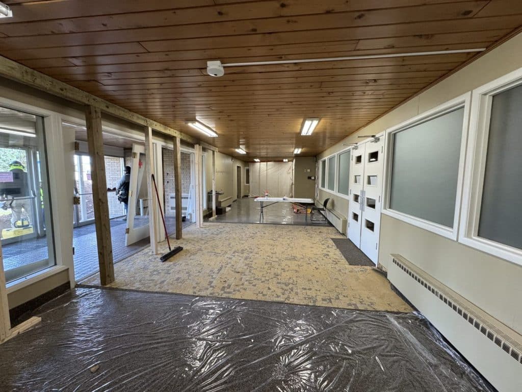 A large indoor hallway under renovation with plastic covering the floor, exposed subfloor, construction tools, and cleaning supplies present.