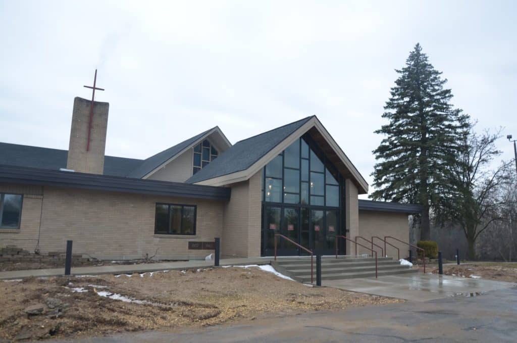 A brick church building with large glass windows, a cross on the chimney, concrete steps, and some patches of snow on the ground, with trees in the background.