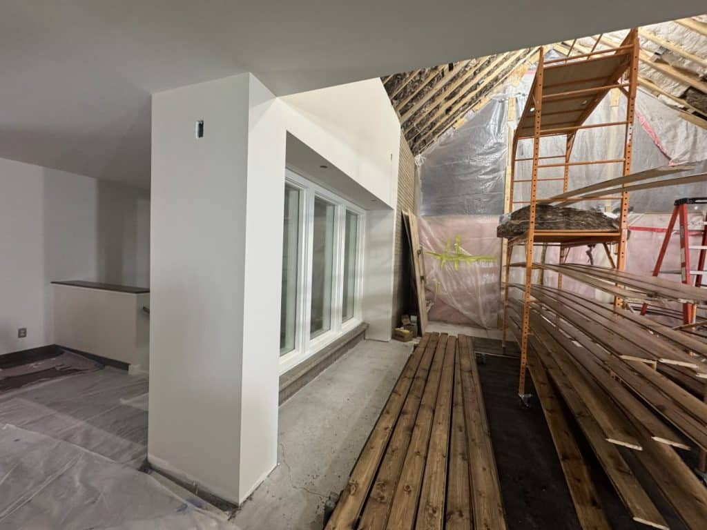 Interior under renovation with exposed rafters, covered flooring, scaffold, stacked wooden planks, and a wall with large windows. Plastic sheets and construction materials are visible.