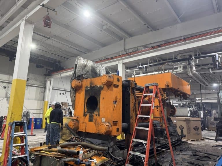 Workers stand near a large, orange industrial machine undergoing maintenance or repair in a spacious factory setting with ladders and tools nearby.