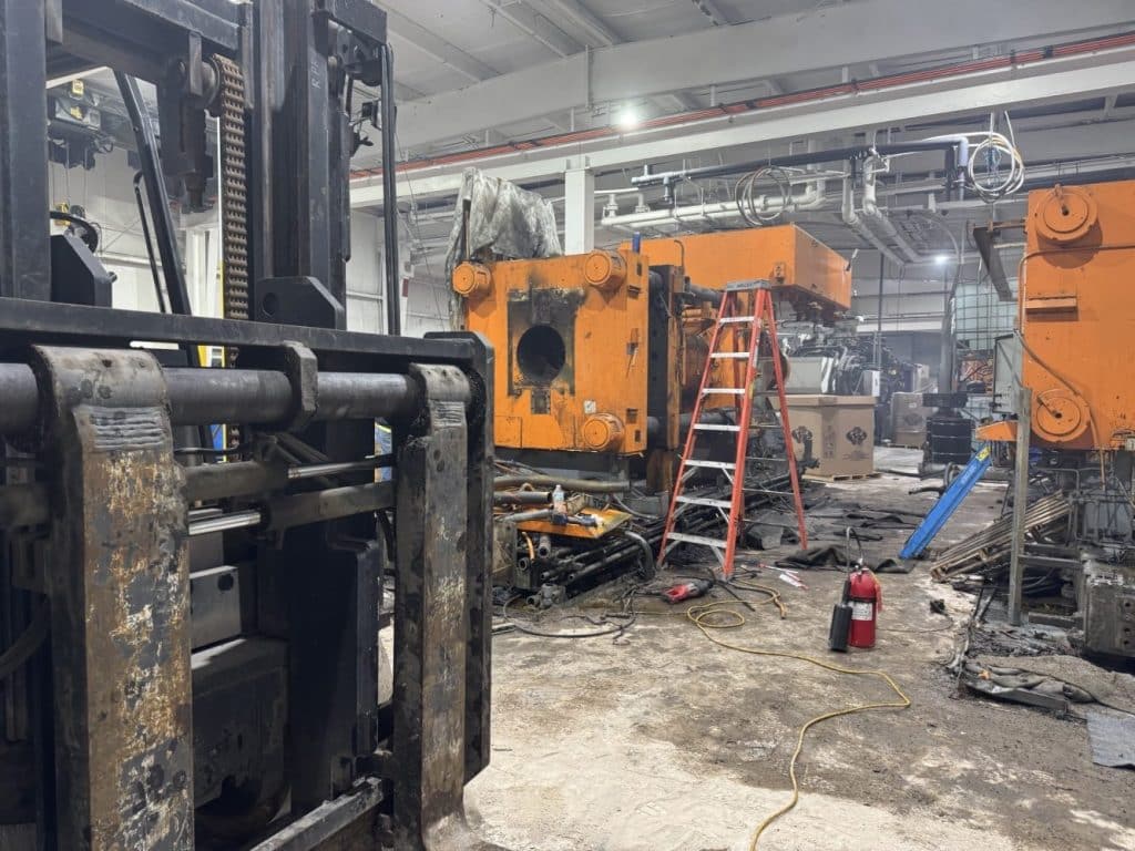 Industrial workshop with orange machinery, a red ladder, a forklift, and scattered tools and debris on the floor; a fire extinguisher is visible in the foreground.
