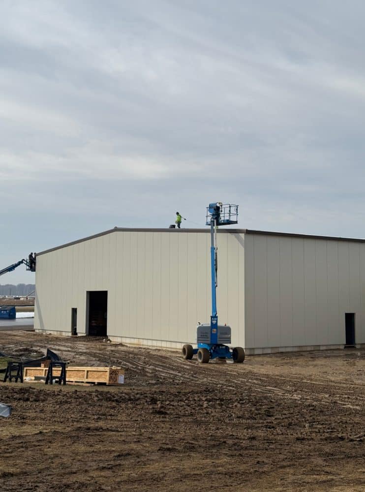 A construction worker stands on the roof of a large industrial building, with a blue boom lift positioned next to the structure on a dirt lot.