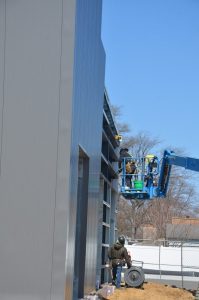 Two construction workers stand on a blue lift while working on the exterior of a metal building; one worker stands on the ground beside building materials.