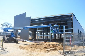 A construction worker operates a blue boom lift beside a partially built metal building, with fencing and construction materials on the sandy ground.