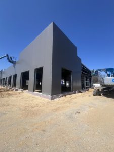 A large, gray industrial building under construction, with sand and construction equipment around the site under a clear blue sky.