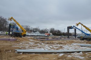 Two construction lifts with workers operate at a snowy, partially built site with scattered materials and overcast sky.