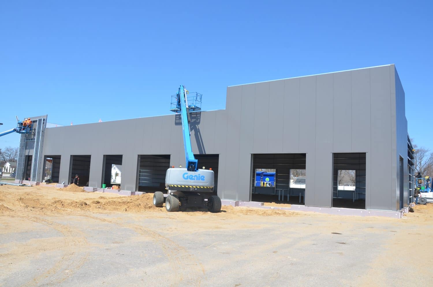 A gray industrial building under construction with open garage bays, a blue Genie lift in front, and workers on site under a clear blue sky.