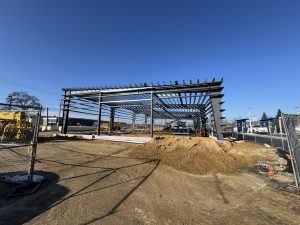 A steel frame structure is under construction on a dirt lot, with construction equipment and materials visible under a clear blue sky.