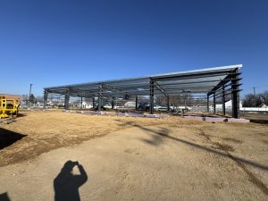 A steel framework of a building under construction on a dirt lot, with construction equipment and clear blue sky in the background.