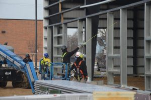 Construction workers wearing safety gear measure and assemble steel beams at a building site with machinery and materials visible.
