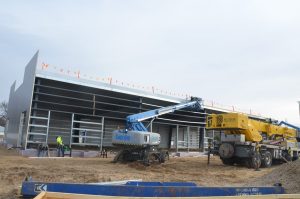 Construction site with workers, a Genie lift, and a crane in front of a partially built metal building structure under an overcast sky.