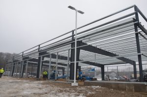 Steel framework of a building under construction on a cloudy day, with workers in safety gear and equipment on site.