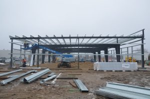 Steel framework of a building under construction on a muddy site, with workers, machinery, and construction materials visible.