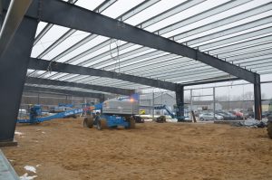 A construction site with a steel frame structure, sandy ground, and a blue Genie S-40 lift. Several vehicles and construction equipment are visible in the background.