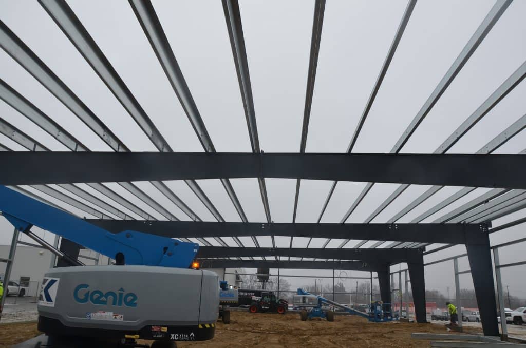Steel beams form the frame of a building under construction, with heavy equipment and workers visible on a cloudy day at the job site.