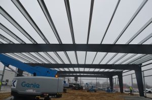 Steel beams form the frame of a building under construction, with heavy equipment and workers visible on a cloudy day at the job site.